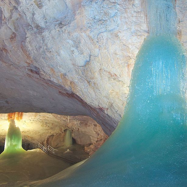 Natural ice formations within a large cave, lit by artificial light, showing smooth, towering ice structures against a rocky cave backdrop.