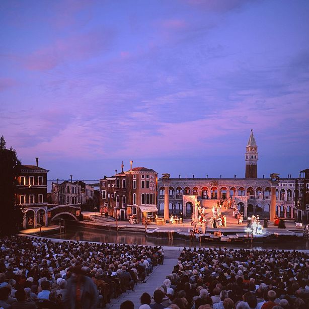 Outdoor theater stage with Venetian architecture backdrop, full audience, and evening lighting. Actors perform under a pink and purple sky.