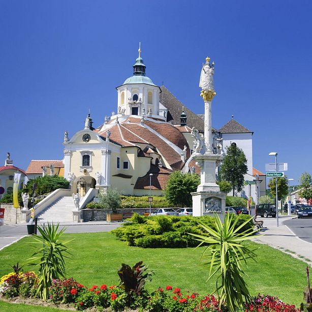 A majestic Baroque church with a large dome and statues, surrounded by lush greenery and a clear blue sky.
