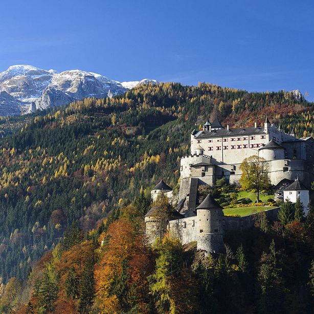 A castle sits on a hill amidst colorful autumn trees, with snow-capped mountains in the background under a clear blue sky.