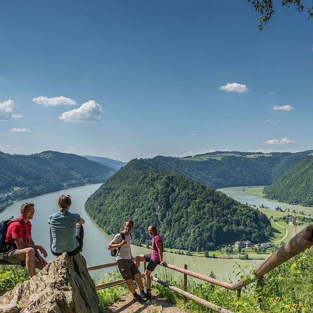 Four hikers sit on a hilltop overlooking a winding river and lush, green hills under a blue sky with scattered clouds.
