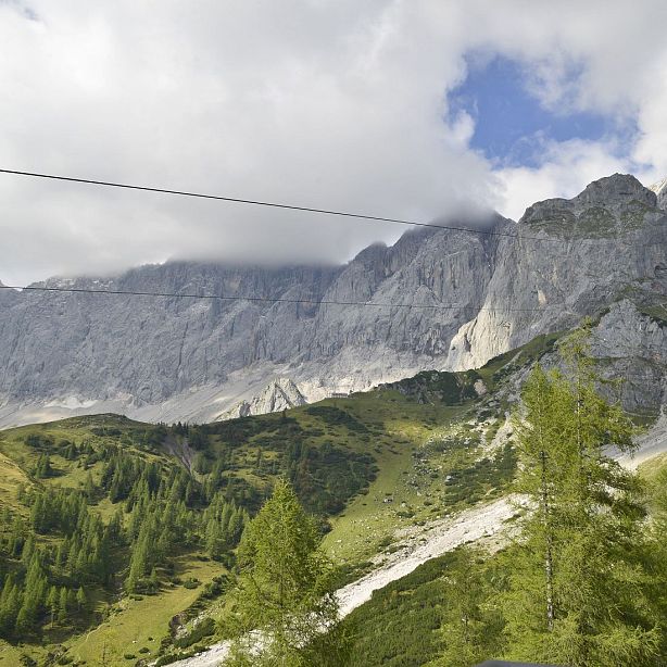 A scenic mountain landscape with towering, cloud-covered peaks and green forested slopes, under a partly cloudy sky. Power lines cross the upper part of the image.
