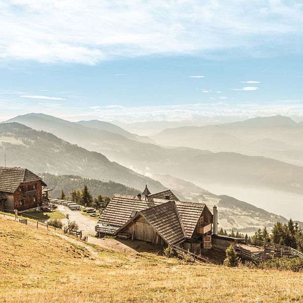 A scenic view of a mountain village with wooden houses, surrounded by misty mountains and a lake, under a clear sky.