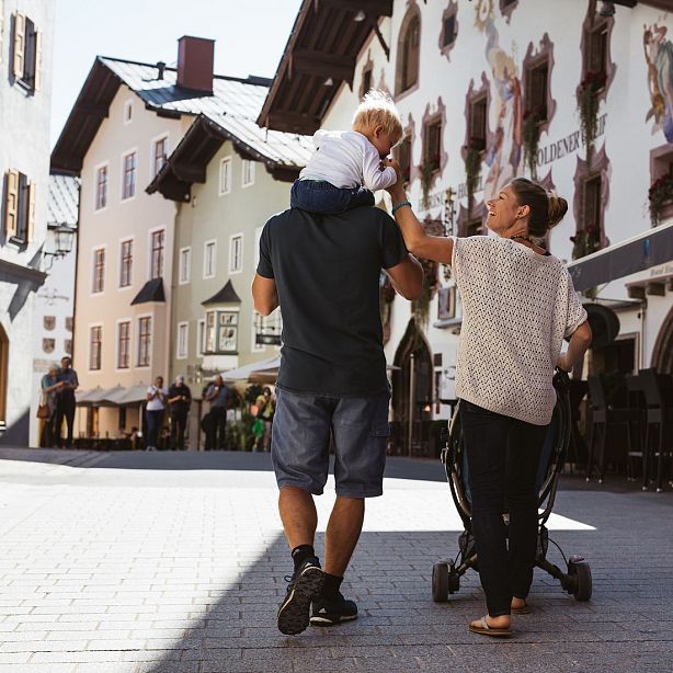 Ein Paar spaziert in einer sonnigen Altstadt: Der Mann trägt ein Kind auf den Schultern, die Frau trägt einen Rucksack. Im Hintergrund sind historische Gebäude.
