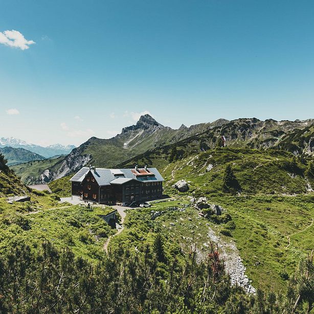 A rustic mountain lodge surrounded by green hills and towering peaks under a clear blue sky, creating a serene and remote natural setting.