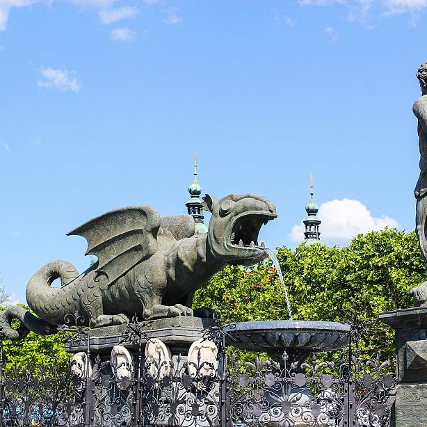 A stone dragon statue with wings and open mouth in a park. Water pours from its mouth into a fountain. Trees and a clear sky in the background.