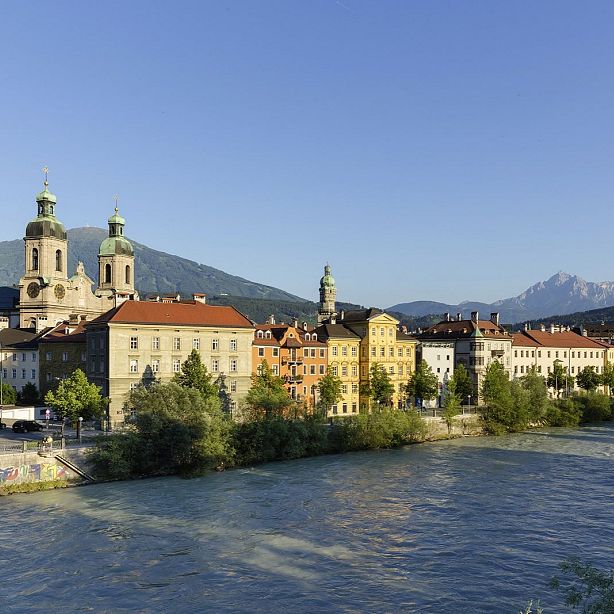 Colorful buildings line the river in Innsbruck, with church towers and mountains in the background, under a clear blue sky.