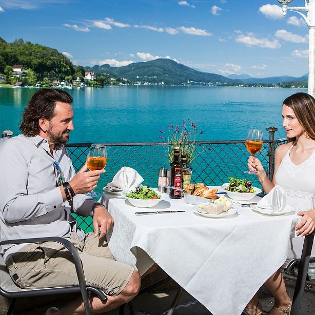 A couple dining outdoors by a lake, sharing drinks and food, with mountains in the background on a sunny day.