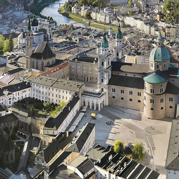Aerial view of a European city square with large, historic buildings, domed towers, and an open plaza. Streets have trees and scattered greenery.