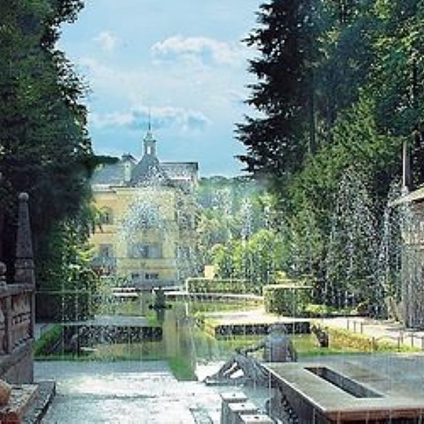 A long garden path with fountains spraying water, lined with trees and hedges, leading toward a distant building under a clear blue sky.