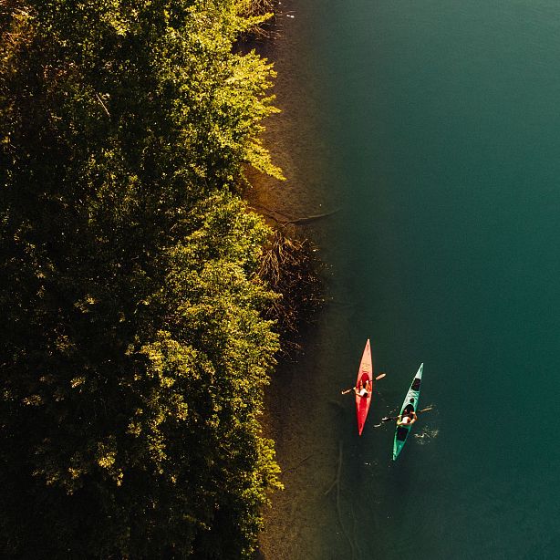Aerial view of two kayaks on a turquoise lake beside lush green foliage. One kayak is red, the other green, each with a person paddling.