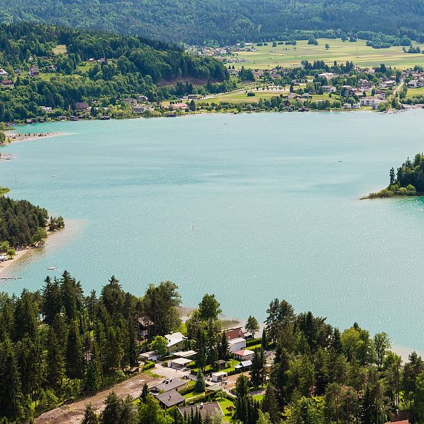 A turquoise lake surrounded by trees and hills with small buildings scattered in the landscape under a clear sky.