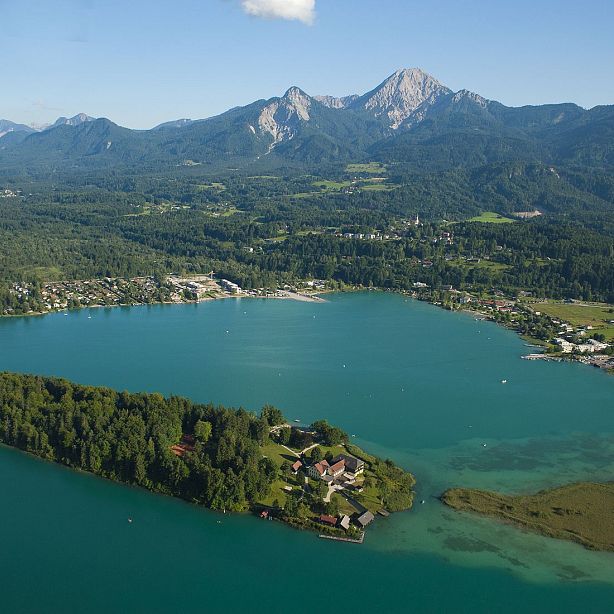 Aerial view of a blue lake surrounded by greenery, with mountains in the distance, small houses along the shore and a peaceful, scenic atmosphere.