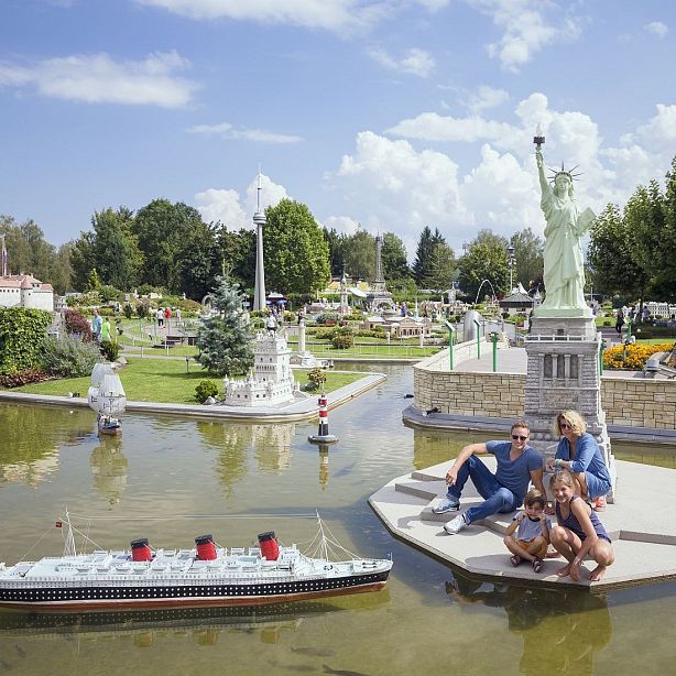 Family at a miniature park with replicas like the Statue of Liberty and a ship, surrounded by trees and a small pond.