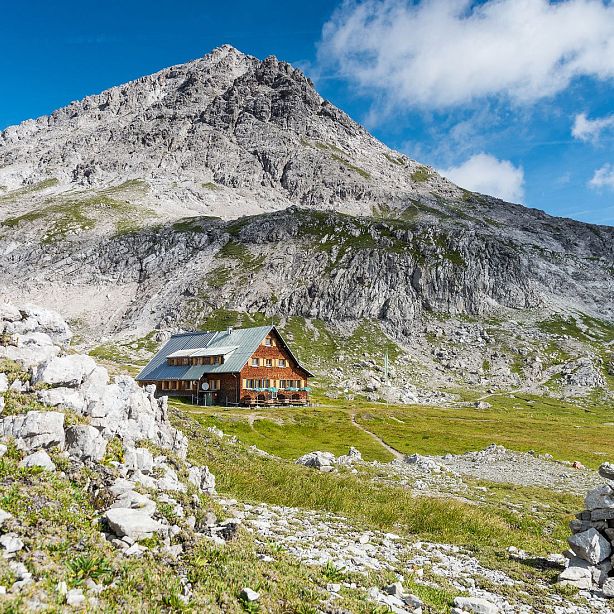 Wooden mountain hut surrounded by rocky terrain and looming mountain under a blue sky with clouds.