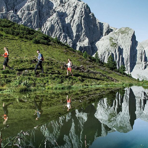 Three people hiking near a lake with mountain reflections and greenery. Majestic rocky mountains stand in the background under a clear sky.