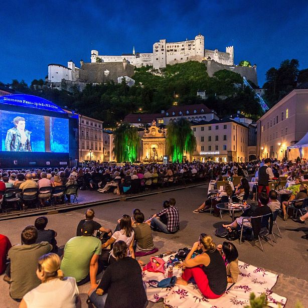 Outdoor nighttime event with people watching a large screen, surrounded by historic buildings and an illuminated fortress in Salzburg, Austria.