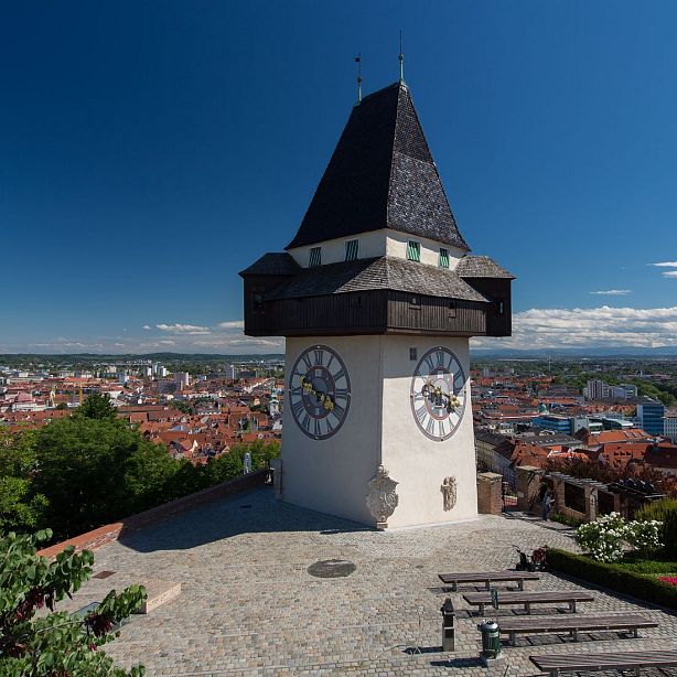 Clock Tower with large clock faces overlooking Graz, Austria, with red rooftops and clear blue sky in background.