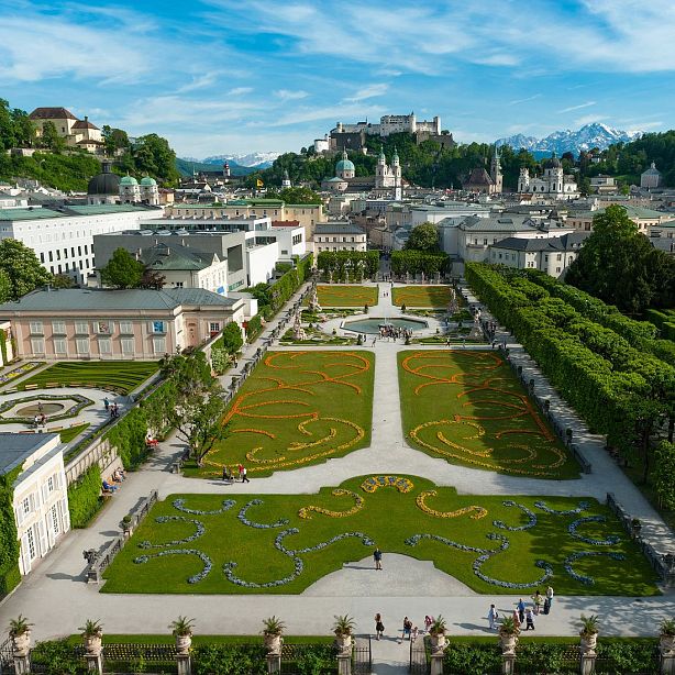 View of Mirabell Gardens in Salzburg with green lawns, winding pathways, sculptures, and a historical city backdrop, set against distant mountains.