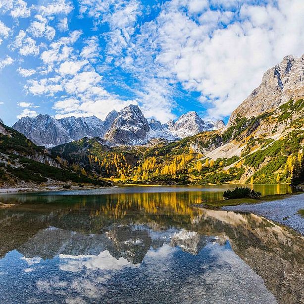 A scenic view of a mountain range with snow peaks, autumn trees in yellow, and a reflective lake beneath a partly cloudy blue sky.