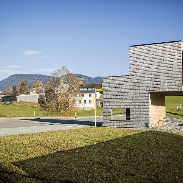 A modern building with wooden cladding in a rural setting, hills in the distance, and a clear blue sky. Green fields and traditional houses surround the scene.
