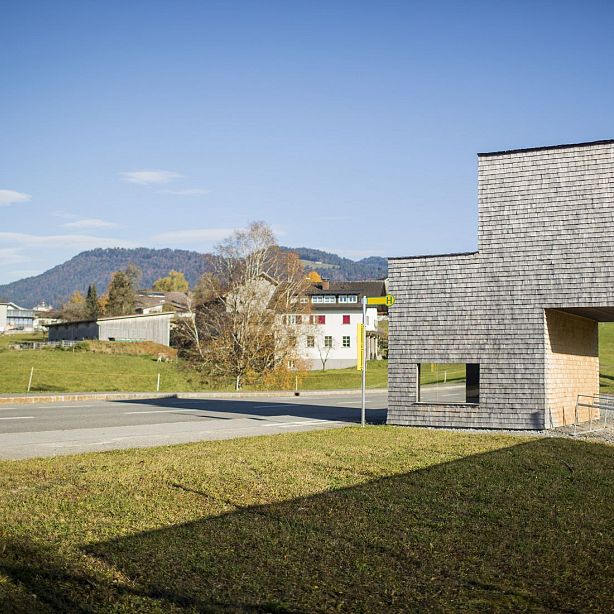 A wooden building sits by a road in a small town, with mountains in the distance under a clear blue sky. Autumn colors are visible in the surrounding trees.