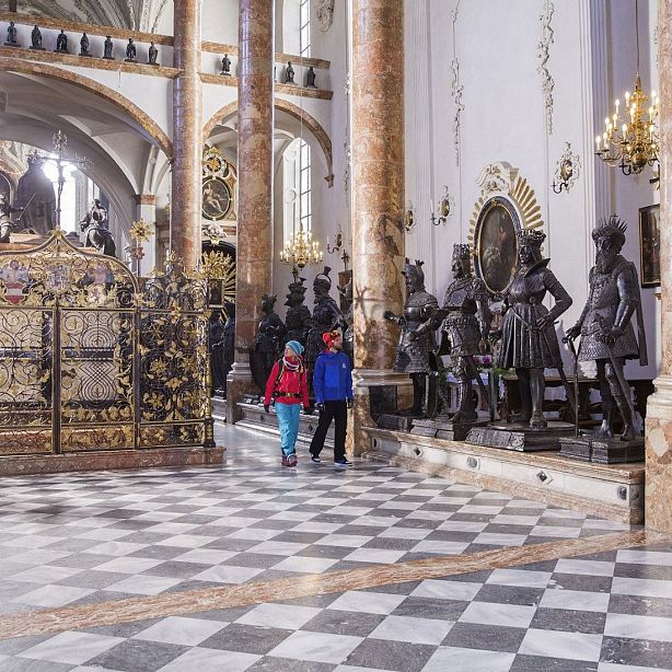 Interior of a historic building with ornate statues, two children walking, checkered floor, chandeliers, and decorative walls. Elegant and grand ambiance.