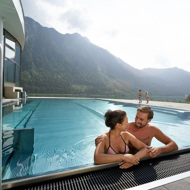 A couple relaxes in an outdoor pool with mountains in the background. The sky is overcast, creating a peaceful and serene atmosphere in the scenic landscape.