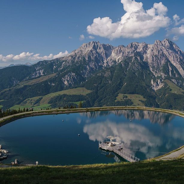 A tranquil lake reflects a majestic mountain range under a partly cloudy sky, with a small dock visible on the water's surface, surrounded by lush greenery.