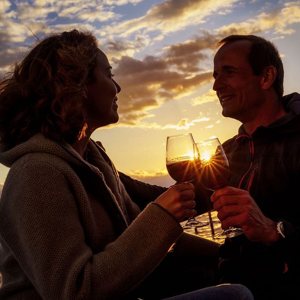 A couple toasting with wine glasses during a warm sunset by the water, silhouettes visible, creating a romantic and serene atmosphere.