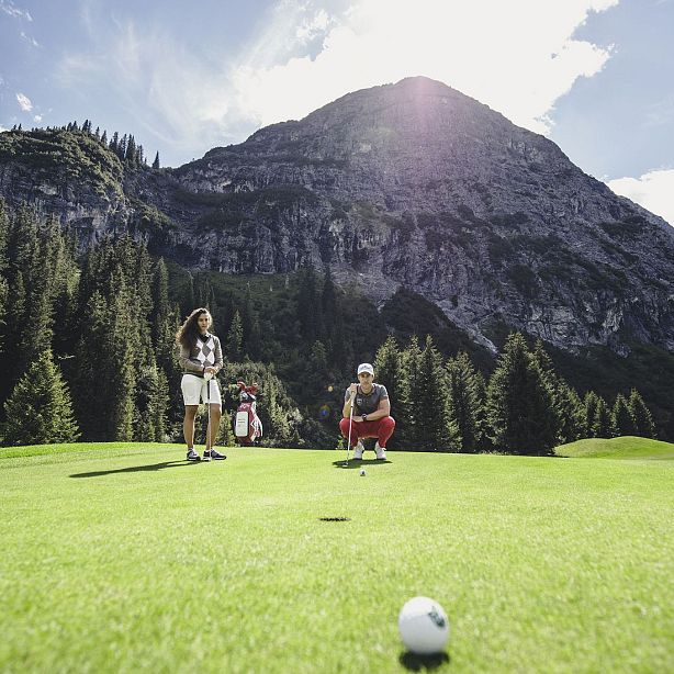 Two individuals playing golf on a lush green course with tall mountains and trees in the background, under a sunny sky.