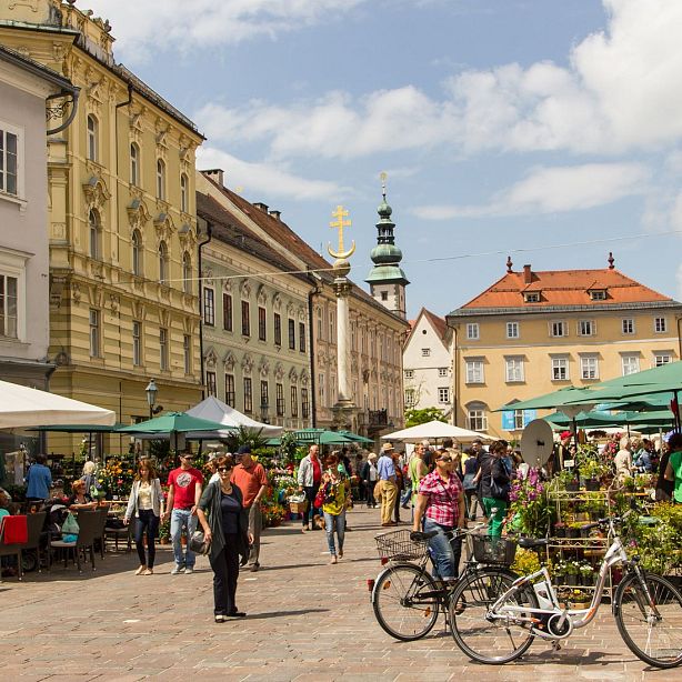 A lively European market square with shoppers and vendors, historic buildings, and green umbrellas covering market stalls on a sunny day.