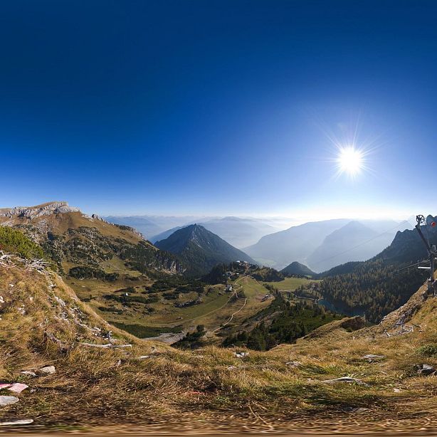 Panoramic view of a mountainous landscape with a bright sun, clear blue sky, grassy slopes, and distant peaks. Ideal for outdoor enthusiasts and nature lovers.