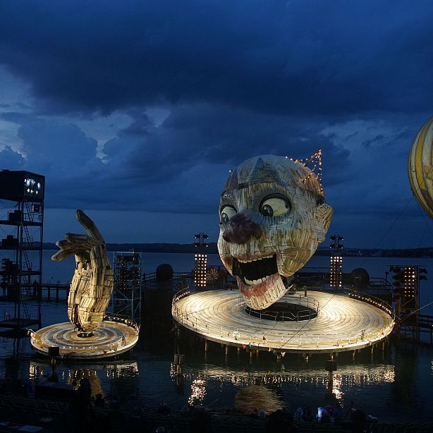 Floating stage with large face sculpture and hand, lit up at night on a lake, against a cloudy sky backdrop.