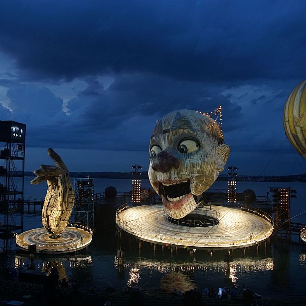 Outdoor theater with giant face and hand sculptures, under a dramatic cloudy sky. Bright lights enhance the surreal, imaginative stage setting.