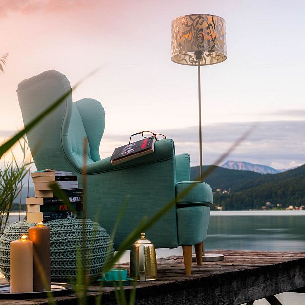 A cozy chair near a stack of books, by a lake with mountains in the background. A calming reading setting with a soft light ambiance.