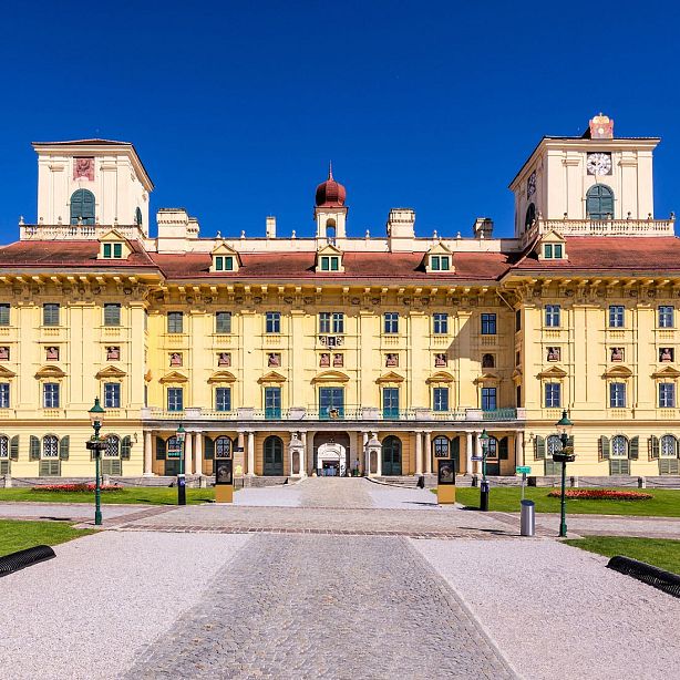 Historic Esterházy Palace with a yellow Baroque facade, two towers, and a central dome, under a clear blue sky, surrounded by a landscaped garden.