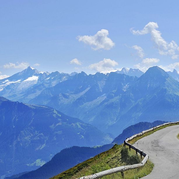 Eine kurvige Bergstraße schlängelt sich entlang eines steilen Abhangs mit einer herrlichen Aussicht auf eine weitläufige Berglandschaft unter blauem Himmel.