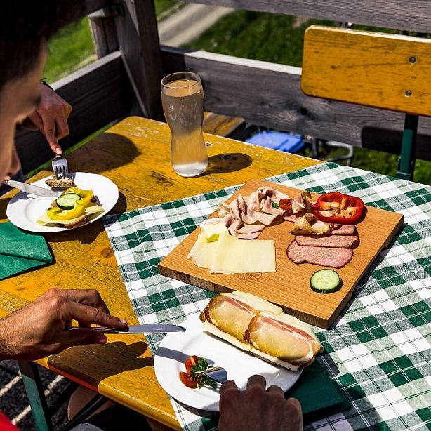 Ein Tisch mit einer Brotzeitplatte: Wurst, Käse, Gemüse und Brot auf einem Holzbrett. Zwei Teller mit ähnlichen Zutaten sind ebenfalls zu sehen.