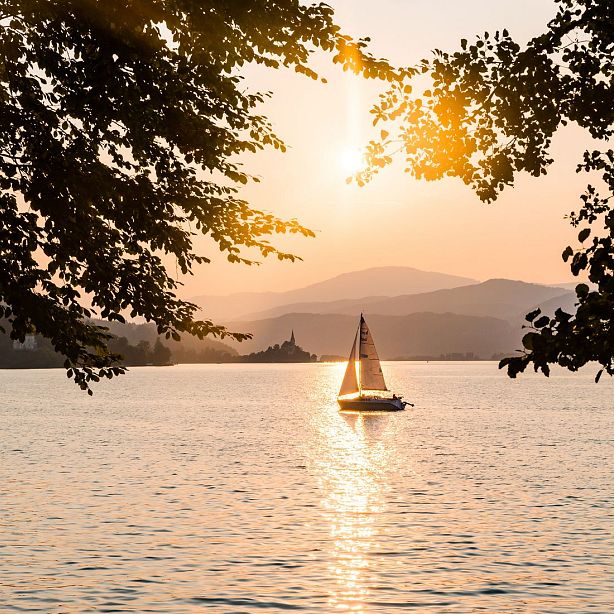 A sailboat on a lake at sunset, surrounded by trees with the sun reflecting on the water. Hills are visible in the background under a golden sky.