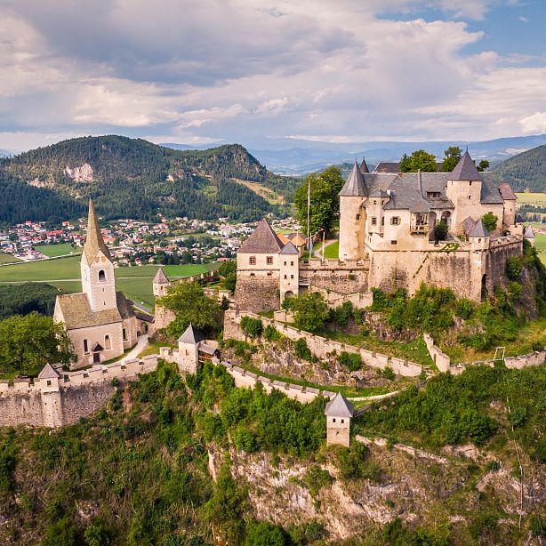Aerial view of Hochosterwitz Castle on hilltop with surrounding greenery, village, and mountains in the background under a clear sky.