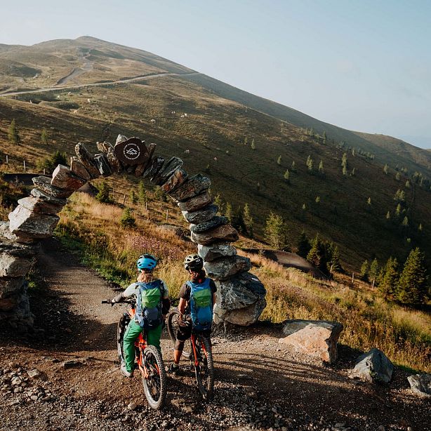 Two mountain bikers stand beneath a stone arch on a sunlit trail, with rolling hills and sparse trees in the background.