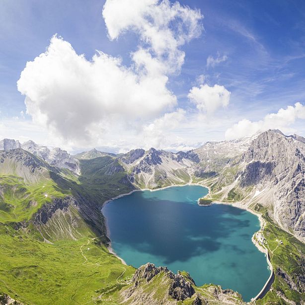 Aerial view of a blue alpine lake surrounded by green mountains and rocky peaks under a blue sky with white clouds.