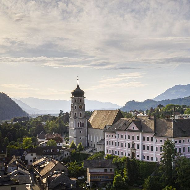 A church with a tall tower in a green valley, surrounded by mountains and cloudy sky, creating a serene landscape.