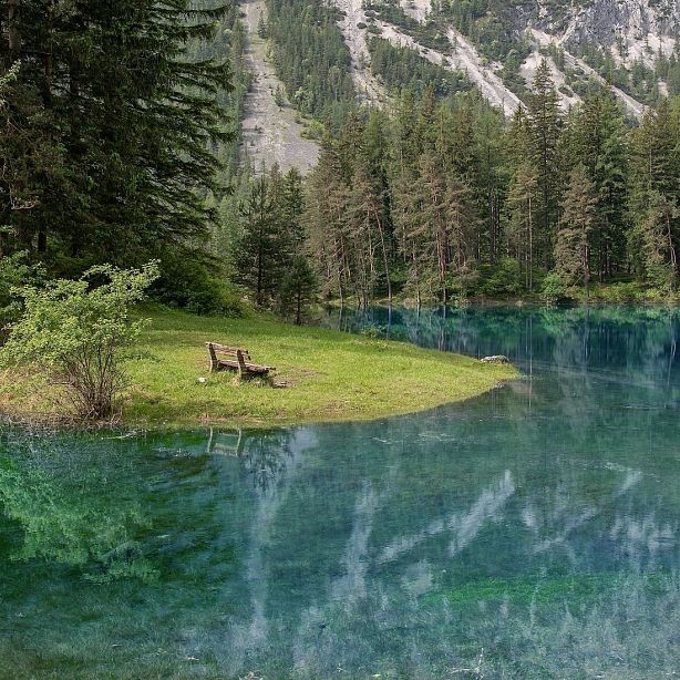 A scenic view of a clear blue lake with a small grassy area and a wooden bench. Trees surround the area, creating a peaceful and natural environment.