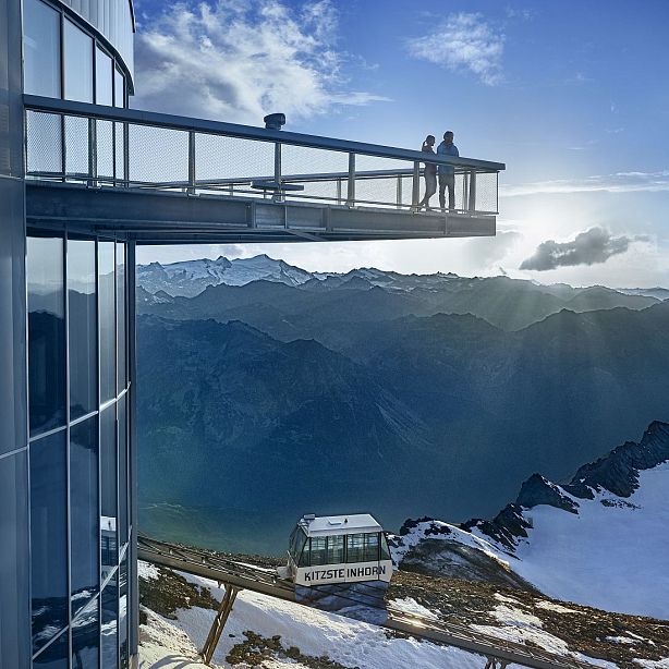 Two people stand on a modern observation deck extending from a mountain building, surrounded by stunning views of snow-capped peaks and valleys under a bright sky.