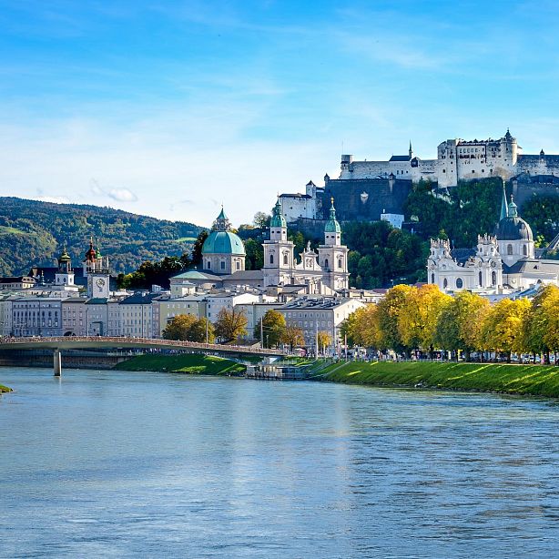 View of Salzburg with Hohensalzburg Fortress on a hill, historical buildings, and the Salzach River in the foreground on a sunny day.