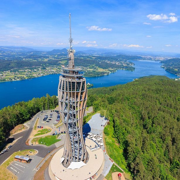 Aerial view of Pyramidenkogel Tower, a spiraling structure beside a parking lot. Surrounded by dense forests, it overlooks a large blue lake and mountains.