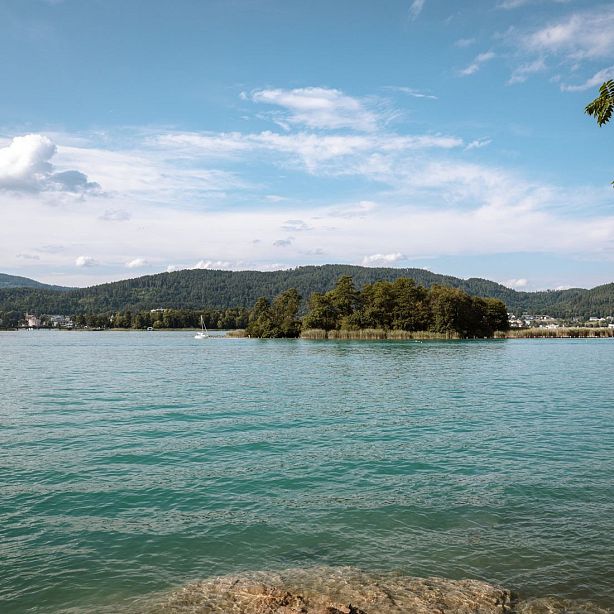 A peaceful lake with blue-green waters, a small island with trees, and distant hills under a partly cloudy sky.