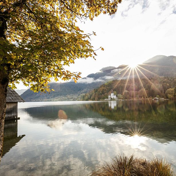 A calm lake with autumn trees, mountains, and sunbeams creating a serene scene. A small boathouse is partly visible on the lake's edge.